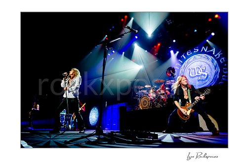 Horizontal color image of David Coverdale, Brian Tichy, and Joel Hoekstra of Whitesnake performing live on stage, with Coverdale at left singing into a microphone, Tichy at center behind the drum kit, and Hoekstra at right playing guitar under vivid blue, red, and white concert lighting