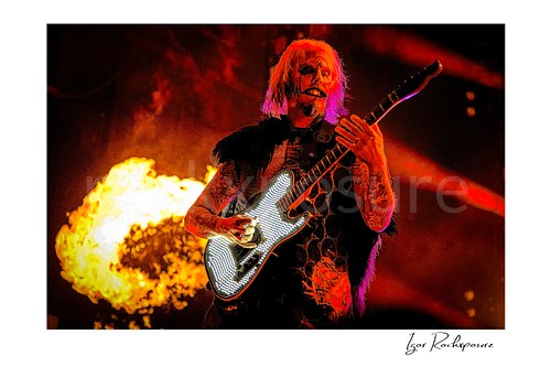Horizontal color image of John 5 performing live on stage with theatrical makeup, a glowing electric guitar, orange fire, and red concert lighting