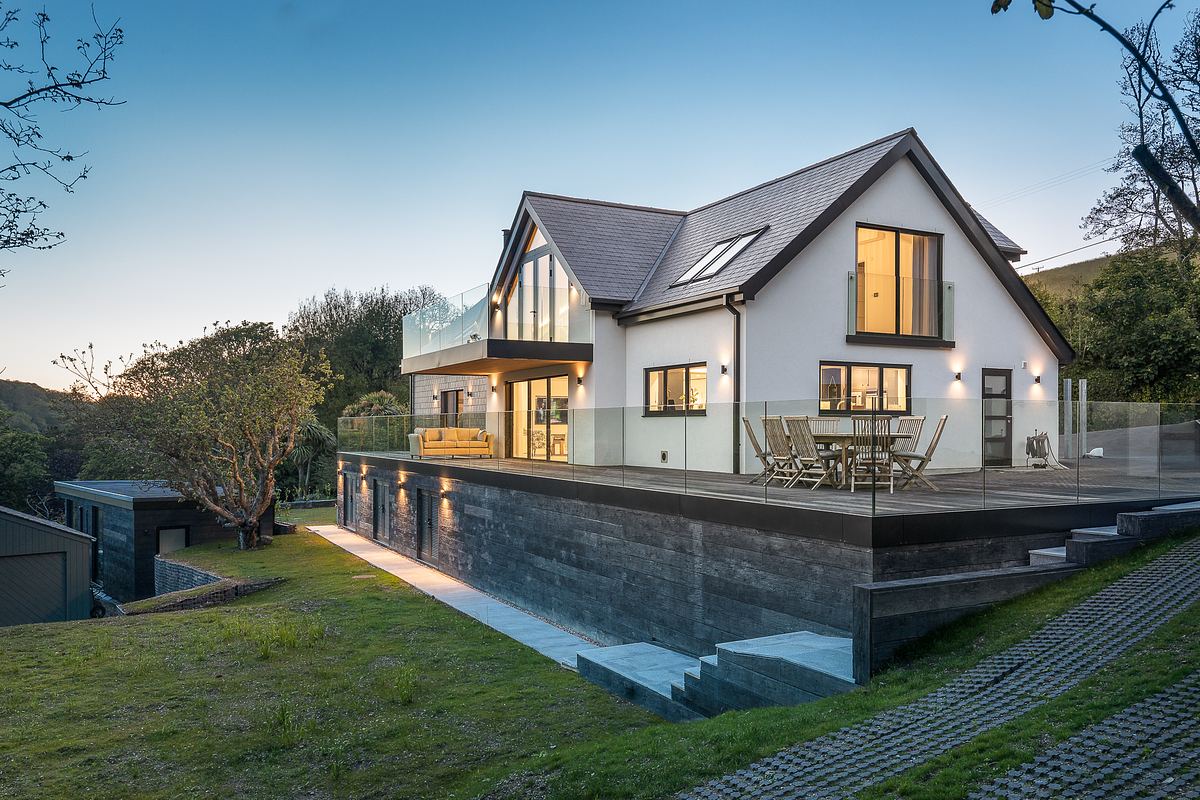 Modern two-story house architect-designed house in Cornwall with large windows, wooden deck, and glass railing, set on a grassy plot at dusk, beautifully captured by a proffessional architectural photographer..
