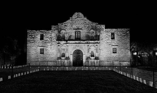 A striking horizontal black and white fine art photograph of the illuminated facade of the Alamo Mission in San Antonio, Texas, captured at night with dramatic shadows.