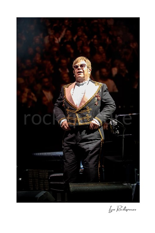 Vertical color image of Elton John standing on stage before a blurred concert crowd in an ornate performance suit