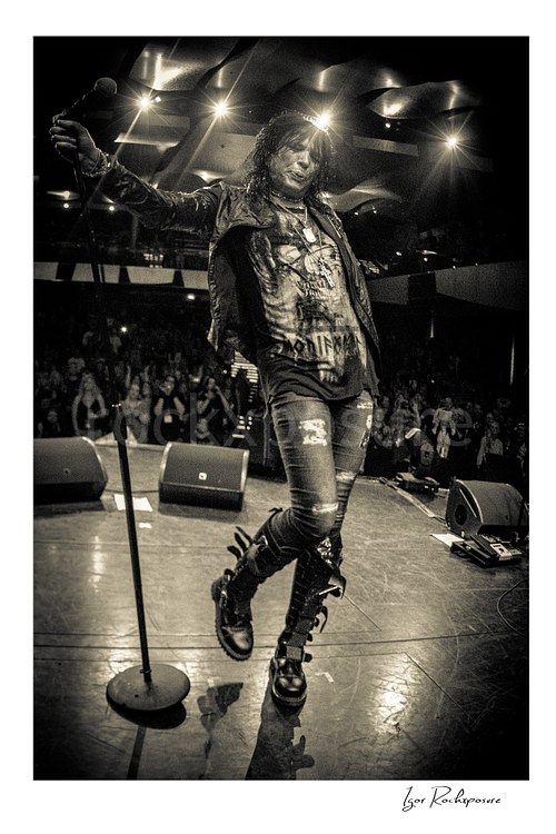 Vertical black and white image of Tom Keifer in a remote camera shot from the drum kit facing forward, walking across the stage with microphone stand in hand as the crowd fills the background