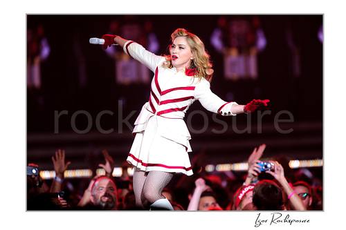 Horizontal color image of Madonna performing in a white and red stage outfit while moving through the crowd with a microphone in hand