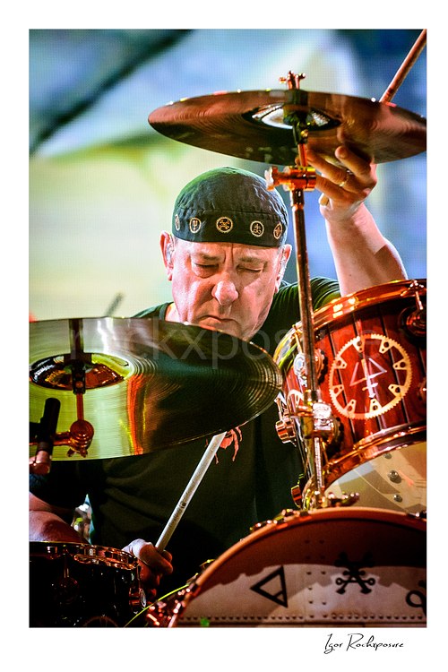 Vertical color image of Neil Peart performing behind his drum kit during a Rush concert, partially framed by cymbals with intense focus and bright stage light
