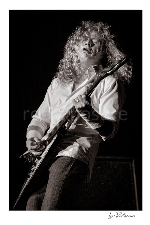 Vertical black and white image of Dave Mustaine of Megadeth playing guitar on stage with flowing curly hair