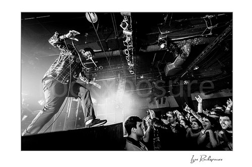 Horizontal black and white image of Chino Moreno of Deftones performing live on stage above the crowd with a handheld microphone, outstretched arm, and fans reaching up