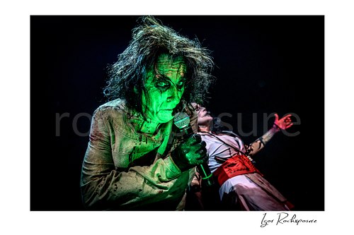 Horizontal color image of Alice Cooper performing live on stage under green lighting with wild hair, horror makeup, and a handheld microphone