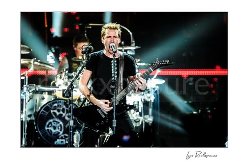 Horizontal color image of Chad Kroeger of Nickelback singing into a microphone while playing guitar with the drummer visible behind him and bright concert beams crossing the stage