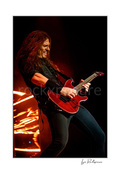 Vertical color image of Kiko Loureiro of Megadeth playing guitar on stage under fiery red lighting