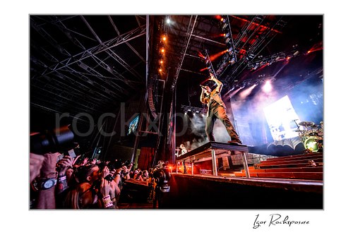 Horizontal color image of Zacky Vengeance of Avenged Sevenfold playing guitar on a stage riser in a dramatic low-angle wide shot with stage smoke and the crowd reaching up from the pit