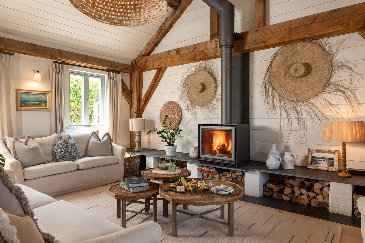 A cozy living room interior with exposed wood beams, a wood-burning stove, two sofas, round coffee tables, decorative baskets on the wall, and neatly stacked firewood.