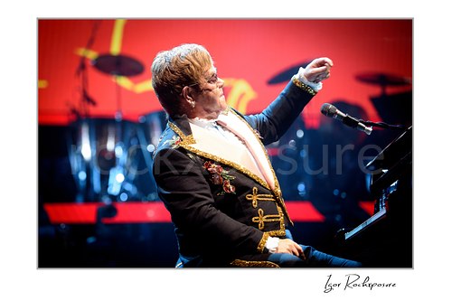 Horizontal color image of Elton John seated at the piano in profile with one arm raised against a vivid red stage backdrop