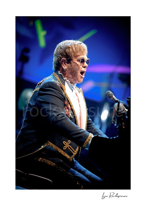 Vertical color image of Elton John singing at the piano in profile under vivid blue stage lighting