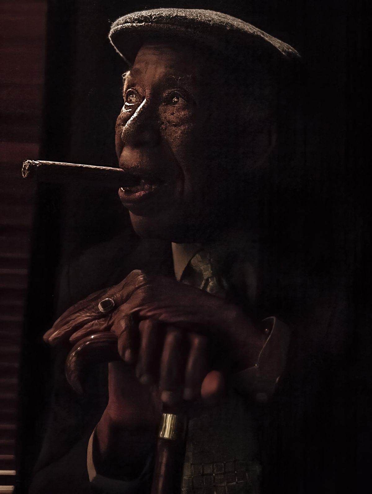 Portrait of Cuban musician Reynaldo. He smokes a cigar and his hands rest on his walkign stick. He is seated.