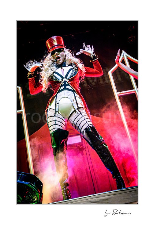 Vertical color image of Maria Brink of In This Moment in a dramatic live performance pose, wearing a red military-style outfit and hat with stage smoke and glowing frame props behind her