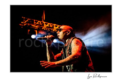 Horizontal color image of M. Shadows of Avenged Sevenfold singing into a microphone with red stage light and a bright beam behind him