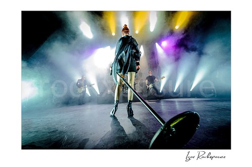 Horizontal color image of Shirley Manson standing on stage during a Garbage concert with microphone stand in the foreground, stage smoke and bright white, yellow and purple lights