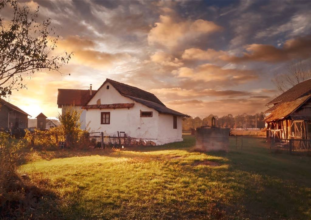 Alt-Tag: Ein traditionelles, altes Bauernhaus mit weißer Fassade in der Region Posavina bei Sonnenuntergang, beleuchtet von warmem Abendlicht unter einem dramatischen Wolkenhimmel.
