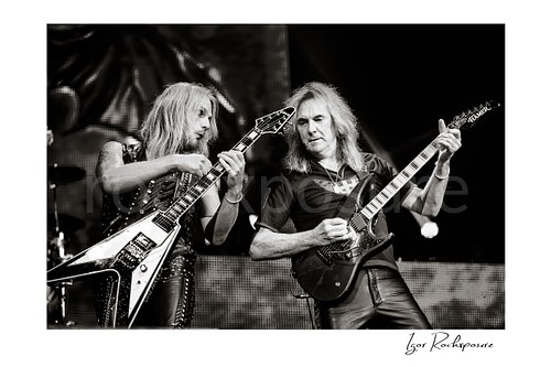 Horizontal black and white image of Richie Faulkner and Glenn Tipton of Judas Priest performing live on stage, with Faulkner at left playing flying V guitar and Tipton at right playing electric guitar