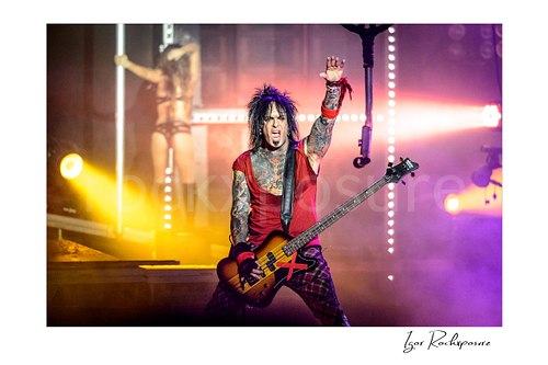 Horizontal color image of Nikki Sixx performing with bass guitar during a Mötley Crüe concert, raising one arm in vivid stage smoke and warm concert light
