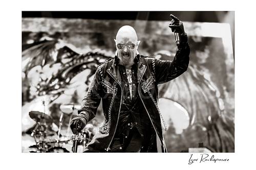 Horizontal black and white image of Rob Halford of Judas Priest performing live on stage, holding a microphone and cane in front of a dramatic LED screen backdrop