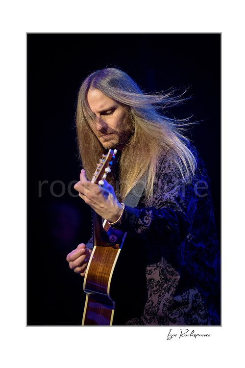 Vertical color image of Jon Davison of Yes playing acoustic guitar onstage with long hair flowing under blue concert light