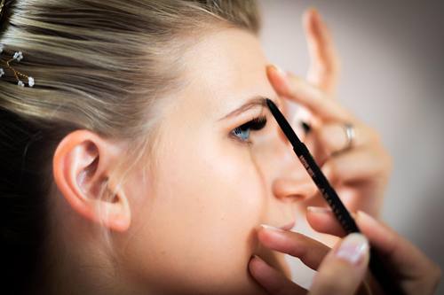 Nahaufnahme im Profil: Eine Braut wird am Hochzeitsmorgen geschminkt, während eine Stylistin die Augenbrauen definiert.