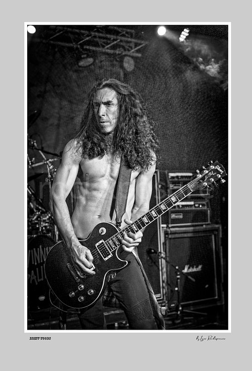 Vertical black and white image of Brent Woods playing guitar shirtless on stage with amplifier stacks behind him