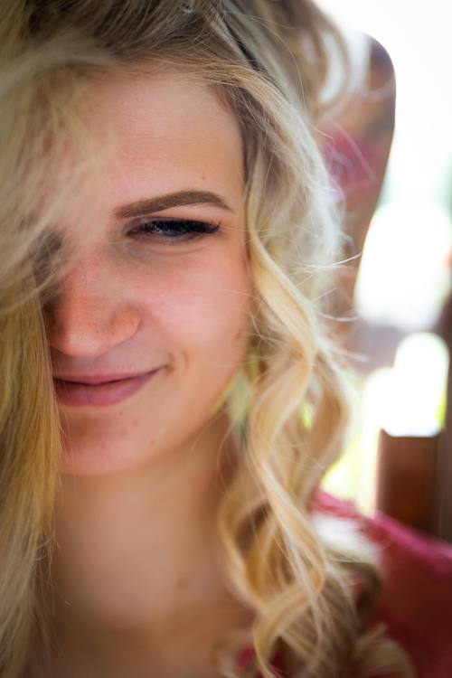 Nahaufnahme einer Braut im Hochformat; eine Gesichtshälfte ist von Haaren bedeckt, während sie mit einem sanften Lächeln frisiert wird.