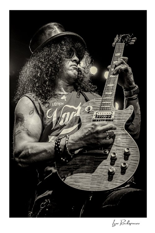 Vertical concert photography of Slash playing electric guitar in black and white photography wearing his signature top hat under dramatic stage lighting