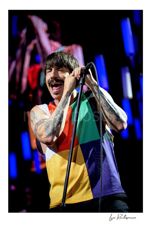 Anthony Kiedis sings with eyes closed and both hands gripping the microphone, tattooed arms framing his multicolor sleeveless top beneath blue LED tubes.