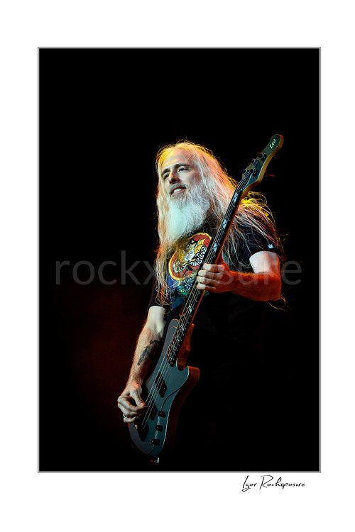 Vertical color image of John Campbell of Lamb of God playing bass on stage with long white beard and warm edge lighting