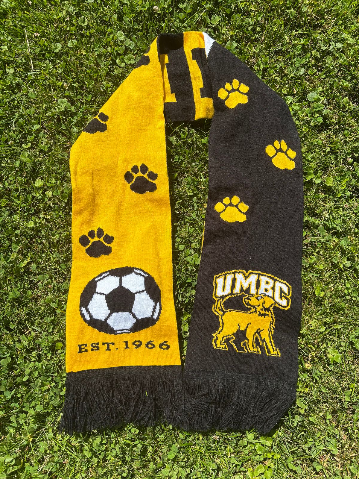 UMBC Black and gold soccer scarf laying on grass