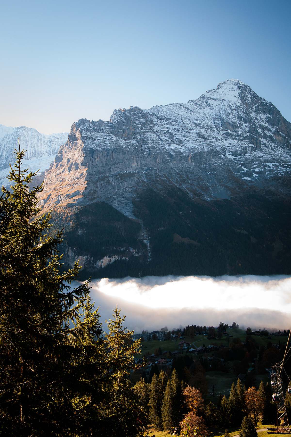Dramatischer Blick auf eine m&auml;chtige Felswand mit Schneefeldern &uuml;ber einem gleissend weissen Wolkenmeer, darunter ein Bergdorf mit Chalets und Herbstb&auml;umen, Gondelkabel im Vordergrund