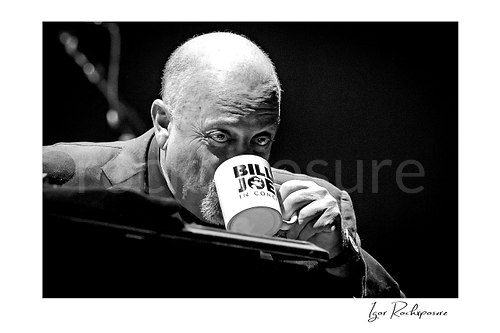 Horizontal black and white image of Billy Joel seated at the piano sipping from a mug in a candid close-up