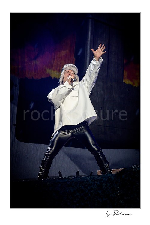 Vertical color image of Bruce Dickinson of Iron Maiden performing live on stage in a white outfit, singing into a microphone with one hand raised