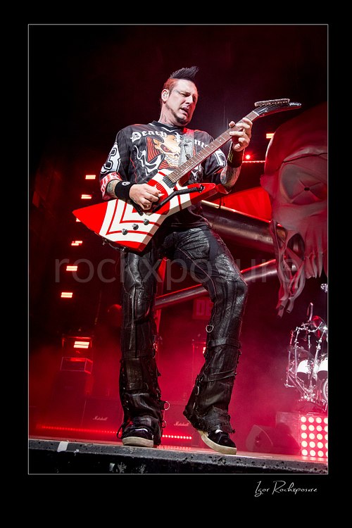 Vertical color image of Zoltan Bathory of Five Finger Death Punch playing guitar onstage under red concert light beside a giant skull stage prop