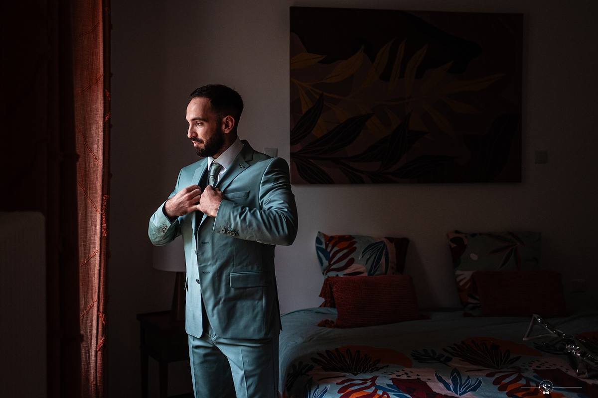 Un mari&eacute; en costume menthe s'ajuste le col de la chemise dans une chambre &eacute;l&eacute;gamment meubl&eacute;e, capturant l'atmosph&egrave;re intime et sereine des pr&eacute;paratifs du mariage c&eacute;l&eacute;br&eacute; sous une tente et dans une prairie &agrave; N&icirc;mes