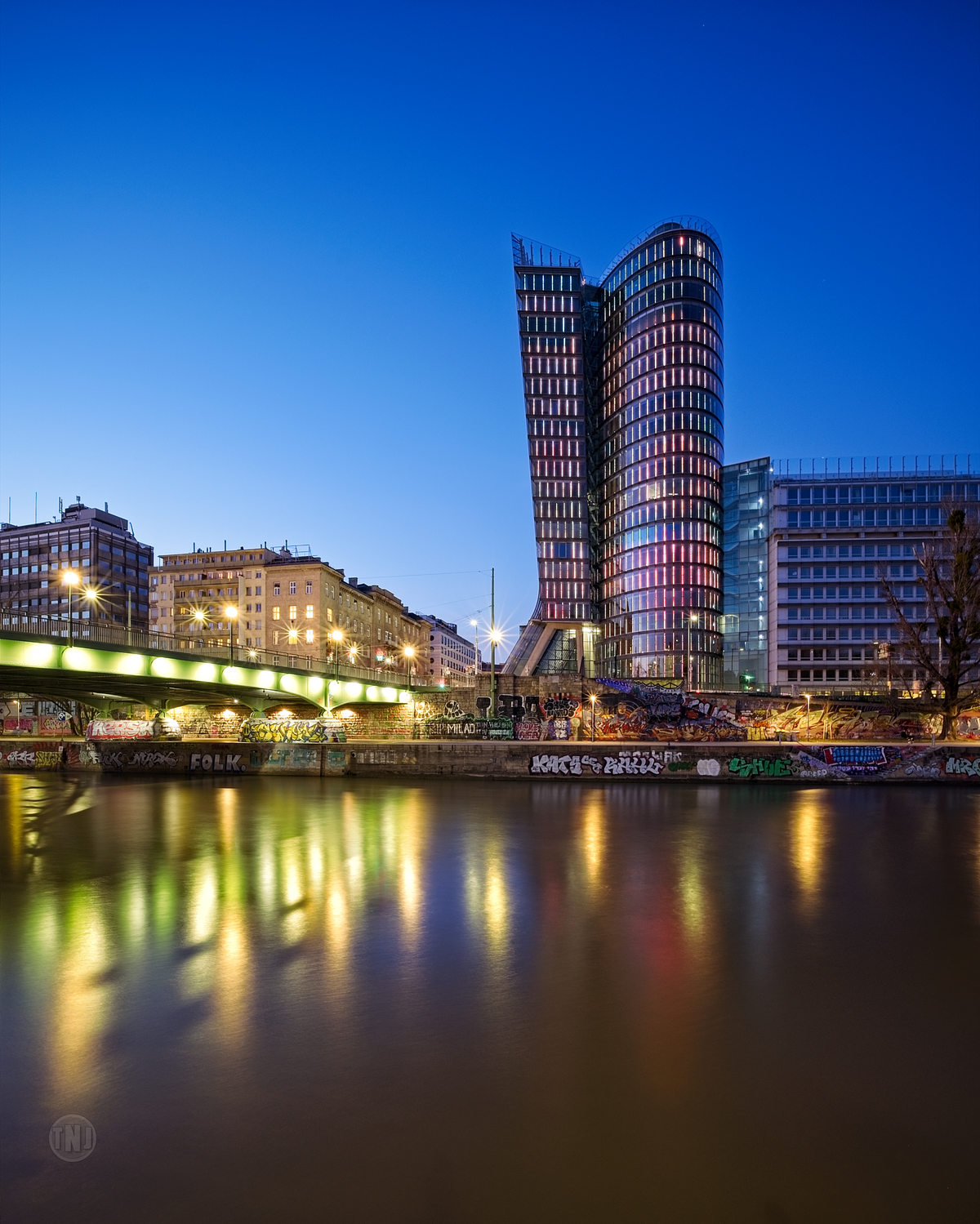 Urbane Nachtaufnahme in Wien: Der beleuchtete Uniqa Tower spiegelt sich im Wasser des Donaukanals neben einer Brücke mit bunten Graffitis.