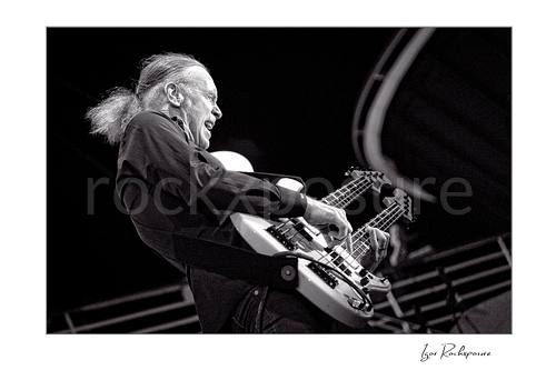 Horizontal black and white image of Billy Sheehan playing his double-neck bass in an energetic side-stage performance shot