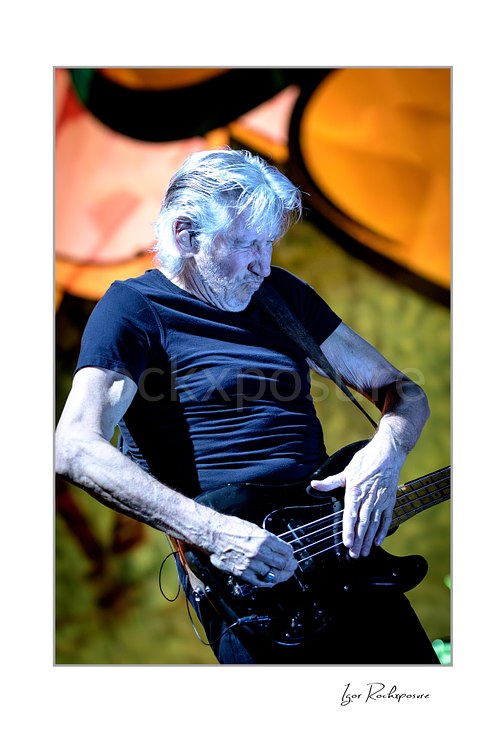 Roger Waters looks down over a black bass guitar with both hands near its body, illuminated by cool light against orange and yellow stage imagery.