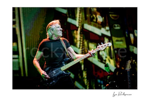 Roger Waters turns his face right while playing black bass under green and warm lighting, backed by a patterned screen and a spare guitar.