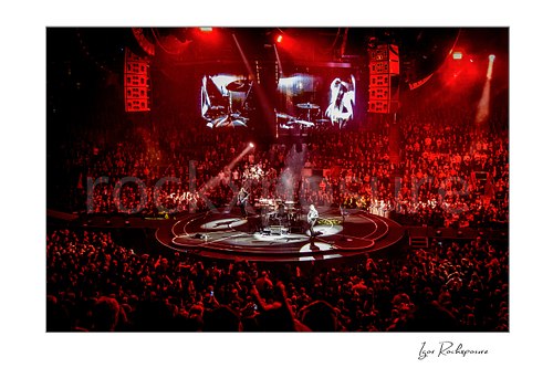 Horizontal color image of Muse performing live on a circular arena stage under vivid red lighting before a packed crowd