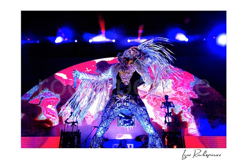 Horizontal color image of Rob Zombie performing live on stage with outstretched arms, flying fringe, vivid blue and pink lighting, and a glowing backdrop screen