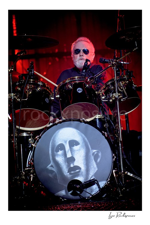 Vertical color image of Roger Taylor of Queen seated behind the drum kit in sunglasses under red concert light