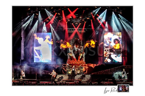 Horizontal color image of Avenged Sevenfold performing on stage in a dramatic full-band wide shot with fire bursts, giant video screens, and vivid red and white concert lights