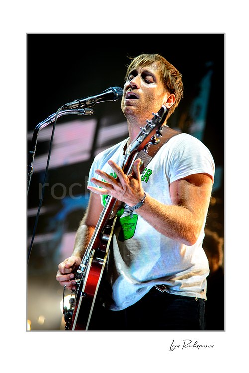 Vertical color image of Dan Auerbach of The Black Keys singing into a microphone while playing guitar in a white T-shirt