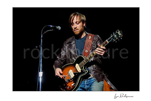Horizontal color image of Dan Auerbach of The Black Keys playing guitar beside a microphone in a dark concert scene