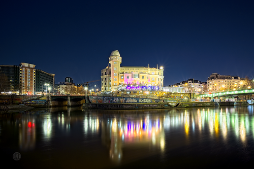 Nachtaufnahme der Urania Sternwarte in Wien vom gegenüberliegenden Ufer des Donaukanals mit bunten Lichtspiegelungen im Wasser.