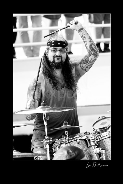 Vertical black and white image of Mike Portnoy at the drum kit with one drumstick raised overhead during a live performance
